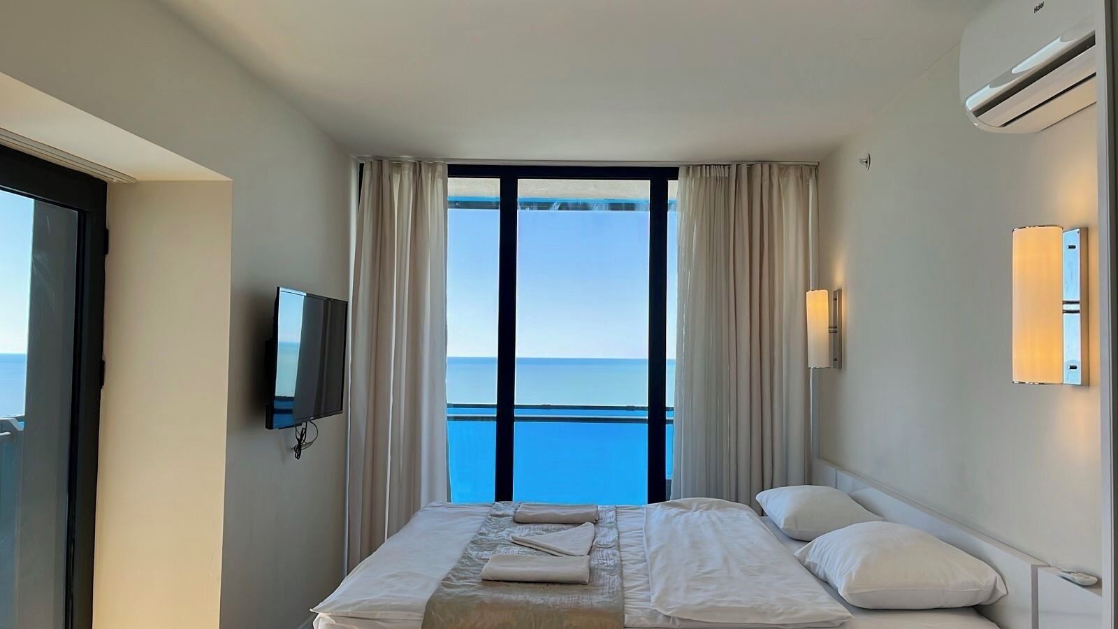 A hotel room with a bed, a TV, a window with curtains, and a view of the sea.