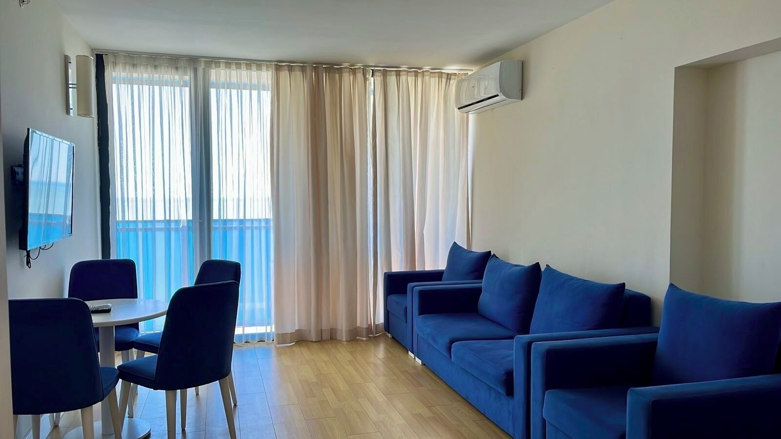 A living room with a sofa set, a dining table with chairs, a flat-screen TV mounted on the wall, and a sliding glass door leading to a balcony.