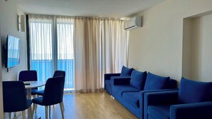 Un salon comprenant un ensemble de canapés, une table à manger avec des chaises, un téléviseur à écran plat fixé au mur et une porte-fenêtre coulissante donnant sur un balcon.