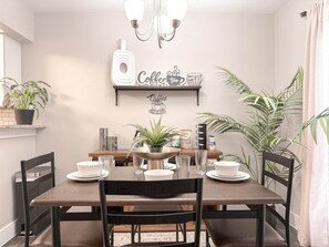 A dining area with a wooden table, chairs, and a chandelier. There are potted plants and a wall-mounted shelf with coffee-themed decor.