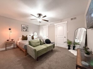 A bedroom with a bed, a green sofa, a ceiling fan, a bedside lamp, a mirror, and a door.