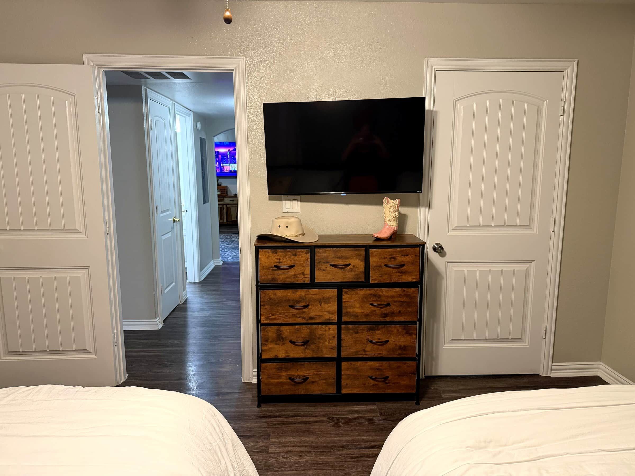 A bedroom with two beds, a dresser with drawers, a television on the wall, and a door leading to another room.