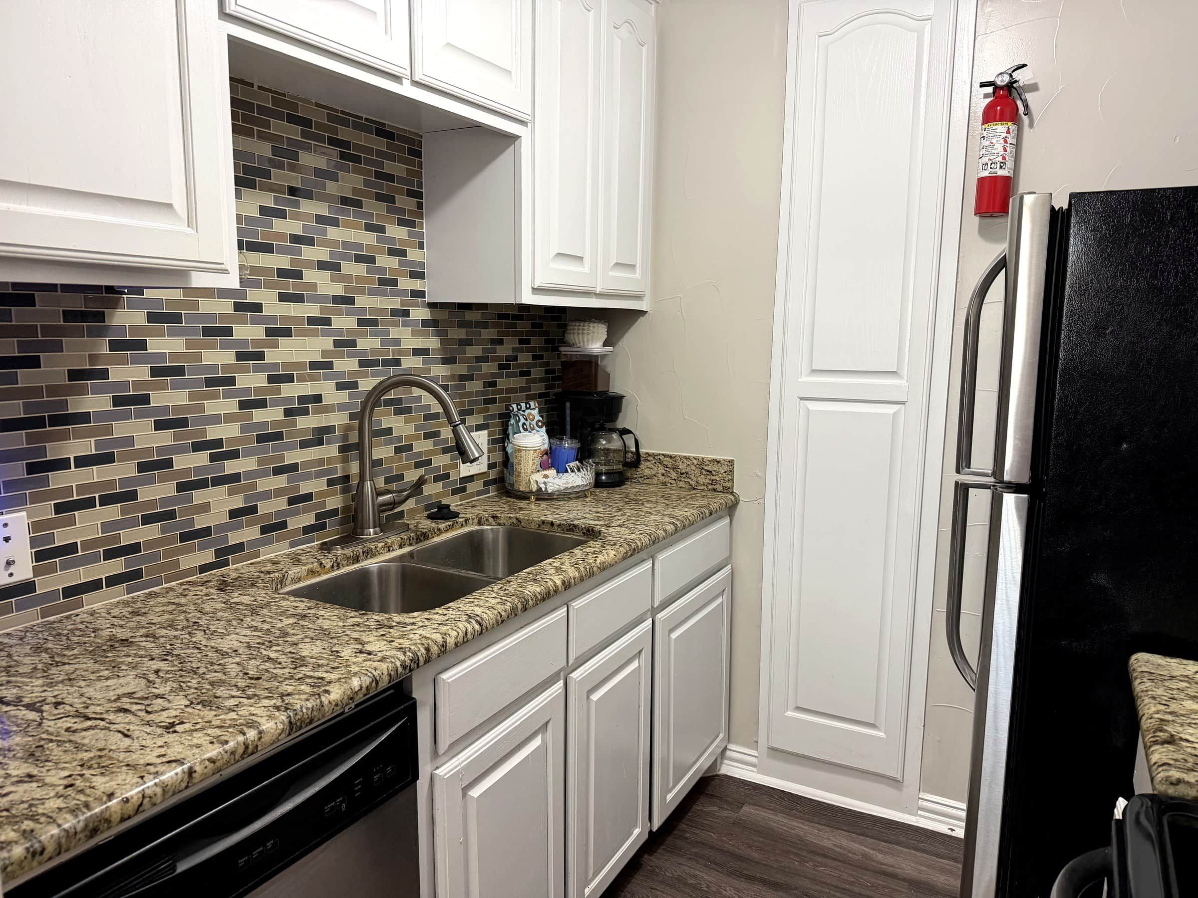 A kitchen with white cabinets, a granite countertop, a stainless steel sink, a coffee maker, and a black refrigerator.
