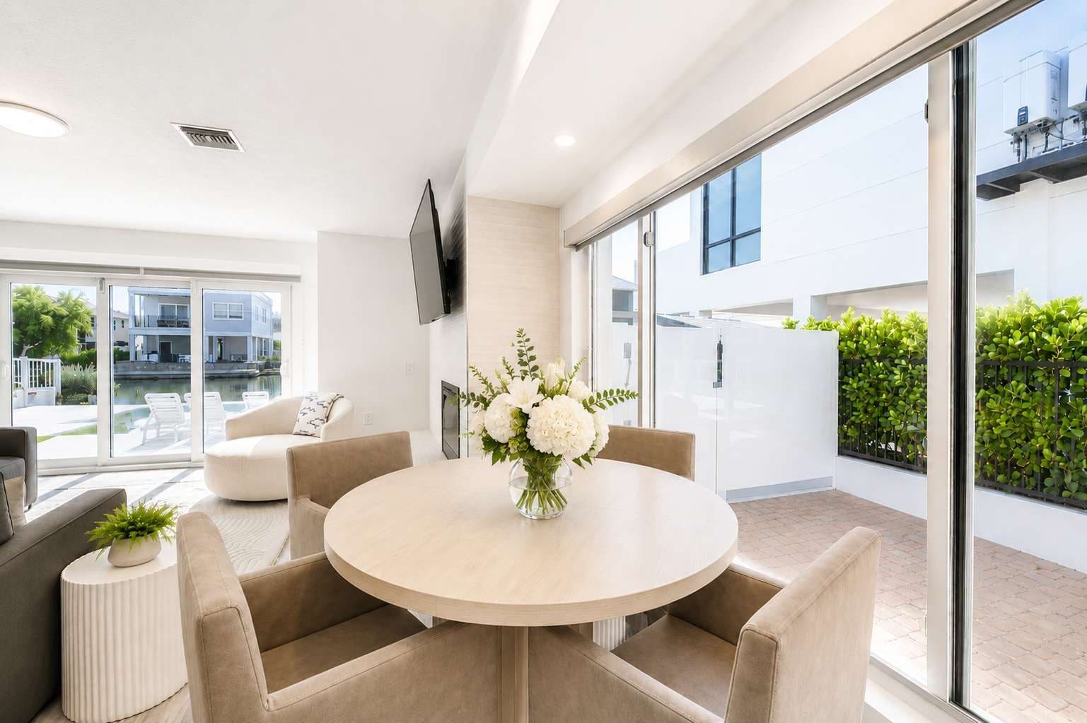 A modern living room with a round table, beige sofas, and a large window offering a view of a pool and buildings.