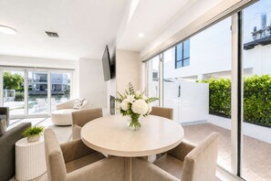 A modern living room with a round table, beige sofas, and a large window offering a view of a pool and buildings. - Vida Isla on Palermo- Heated Pool- Sleeps 10 (Fort Myers Beach)