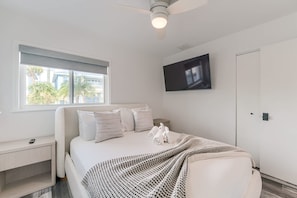 A modern bedroom with a bed, a TV mounted on the wall, and a window with a view of palm trees. - Vida Isla on Palermo- Heated Pool- Sleeps 10 (Fort Myers Beach)
