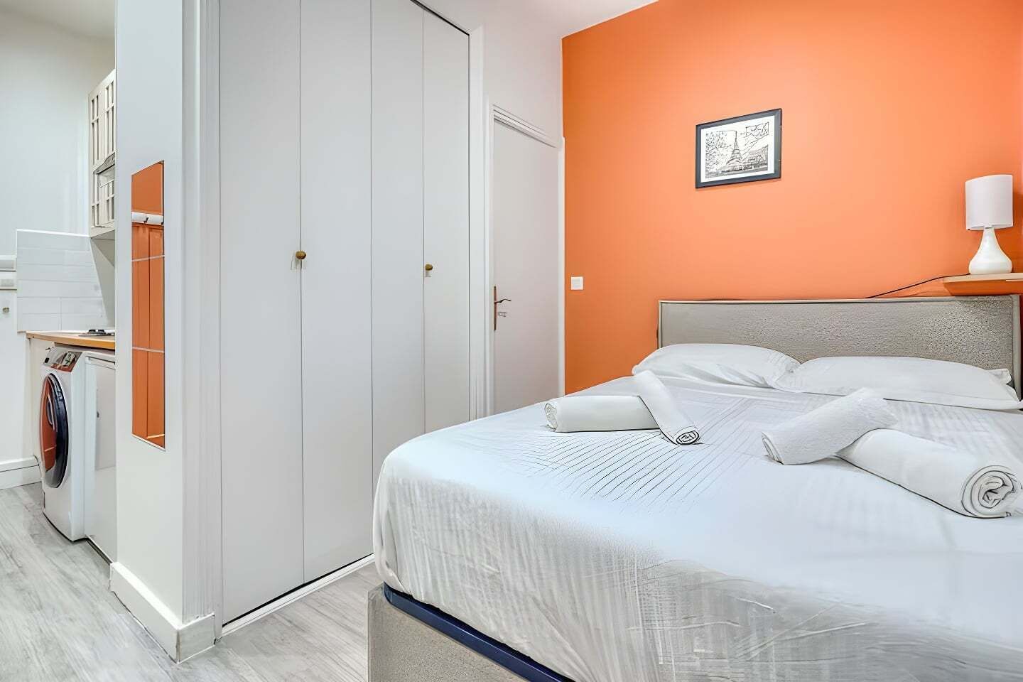 A hotel room with a bed, a wardrobe, and a small table with a lamp.