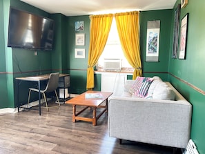 A room with a flat-screen TV, a desk, a chair, a sofa, and a window with yellow curtains. - Cozy & Quiet 1 Bed/1 Bath Rittenhouse Condo (Philadelphia)