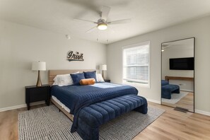 Une chambre à coucher comprenant un lit, des tables de chevet, un ventilateur de plafond, une fenêtre avec des stores, un miroir et un téléviseur fixé au mur.