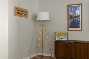 A room with a floor lamp, a cabinet, a framed picture, and a sign on the wall. - The Irish Landing | 2BR/2BA | Private Balcony (South Bend)