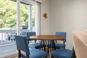 Un espace repas avec une table ronde et six chaises, un plancher en bois, une rambarde blanche et une vue sur les arbres et les voitures garées à l’extérieur.