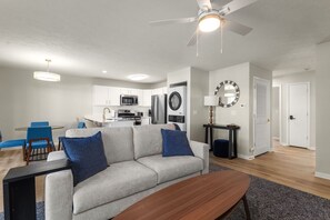 A modern living room with a grey sofa, blue cushions, a wooden coffee table, and a ceiling fan. - The Irish Landing | 2BR/2BA | Private Balcony (South Bend)