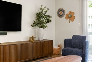A living room with a flat-screen TV, a wooden cabinet, a blue armchair, a white vase with a plant, and two wooden wall decorations. - The Irish Landing | 2BR/2BA | Private Balcony (South Bend)