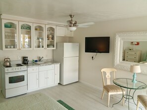 A compact kitchen with white cabinets, a stove, sink, and refrigerator. There's a ceiling fan, a TV mounted on the wall, and a small round table with a chair. - Unit 117 - Comfy Island Treasure (Ground Floor) (Treasure Island)