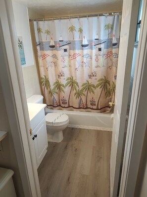 A bathroom with a toilet, a shower curtain with a beach theme, and a wooden floor. - 121 - Beach Bum Break House (2nd Floor) (Treasure Island)