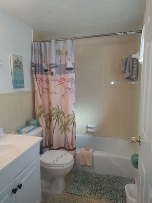 Ein Badezimmer mit Toilette, einer Badewanne, einer Duschkabine mit Strandmotiv, einem Handtuchhalter und einer kleinen Ablage für Toilettenartikel.