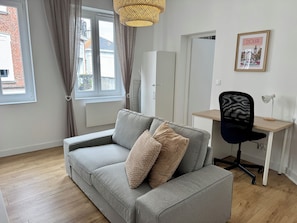 Een moderne woonkamer met een grijze bank, een zwarte bureaustoel, een houten bureau en een fotolijst aan de muur.