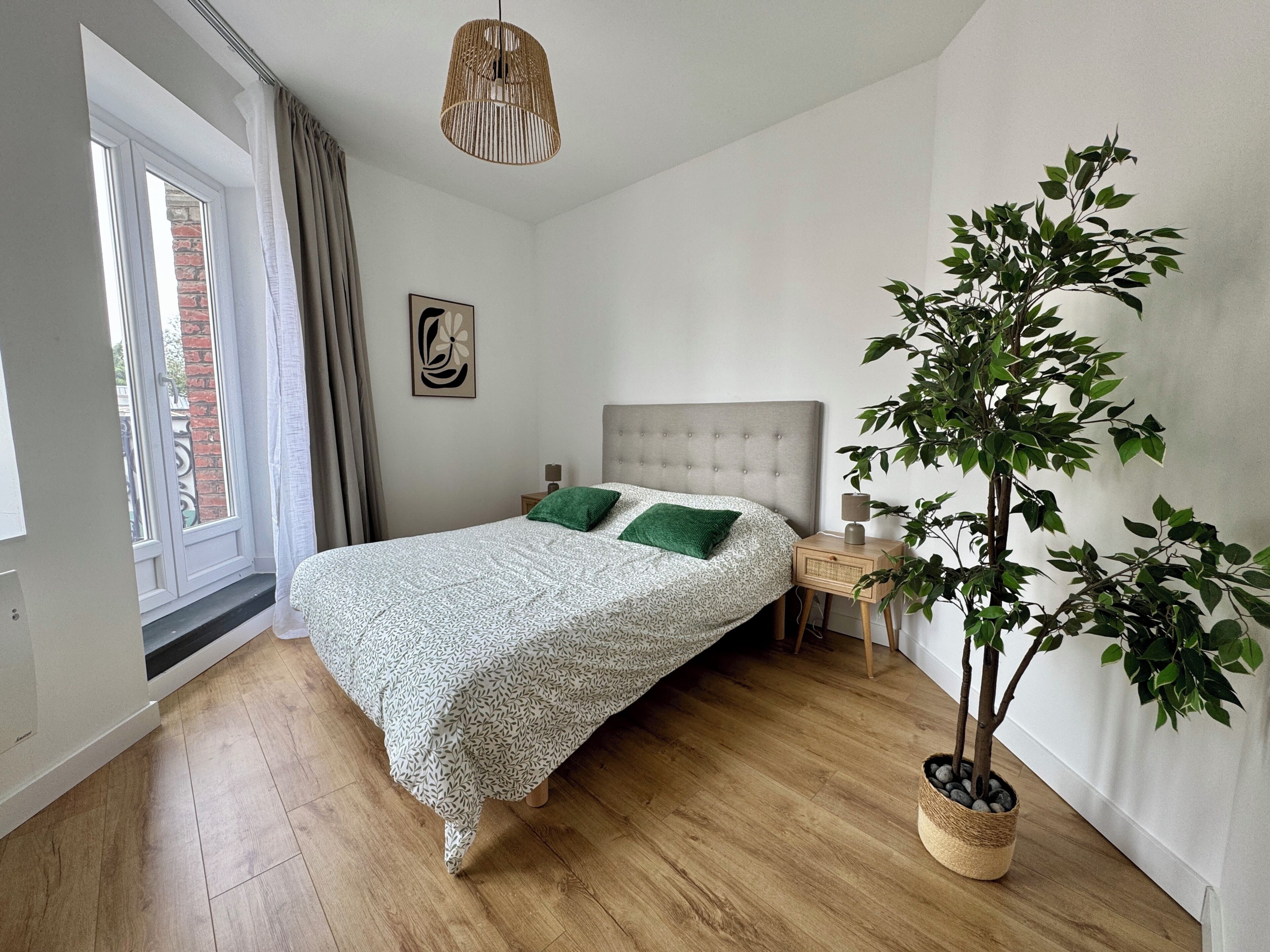 Una camera da letto con un letto, un comodino, una lampada, una lampada a sospensione, una finestra con tende e una pianta in vaso.