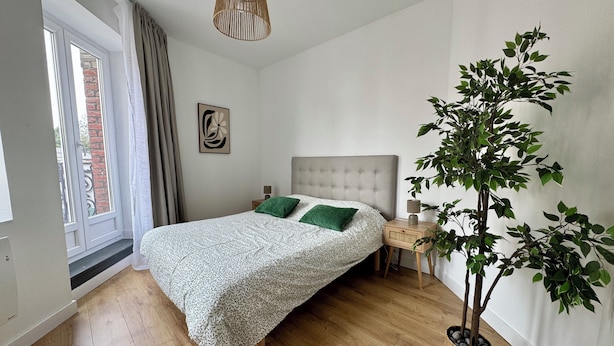 Una camera da letto con un letto, un comodino, una lampada, una lampada a sospensione, una finestra con tende e una pianta in vaso.