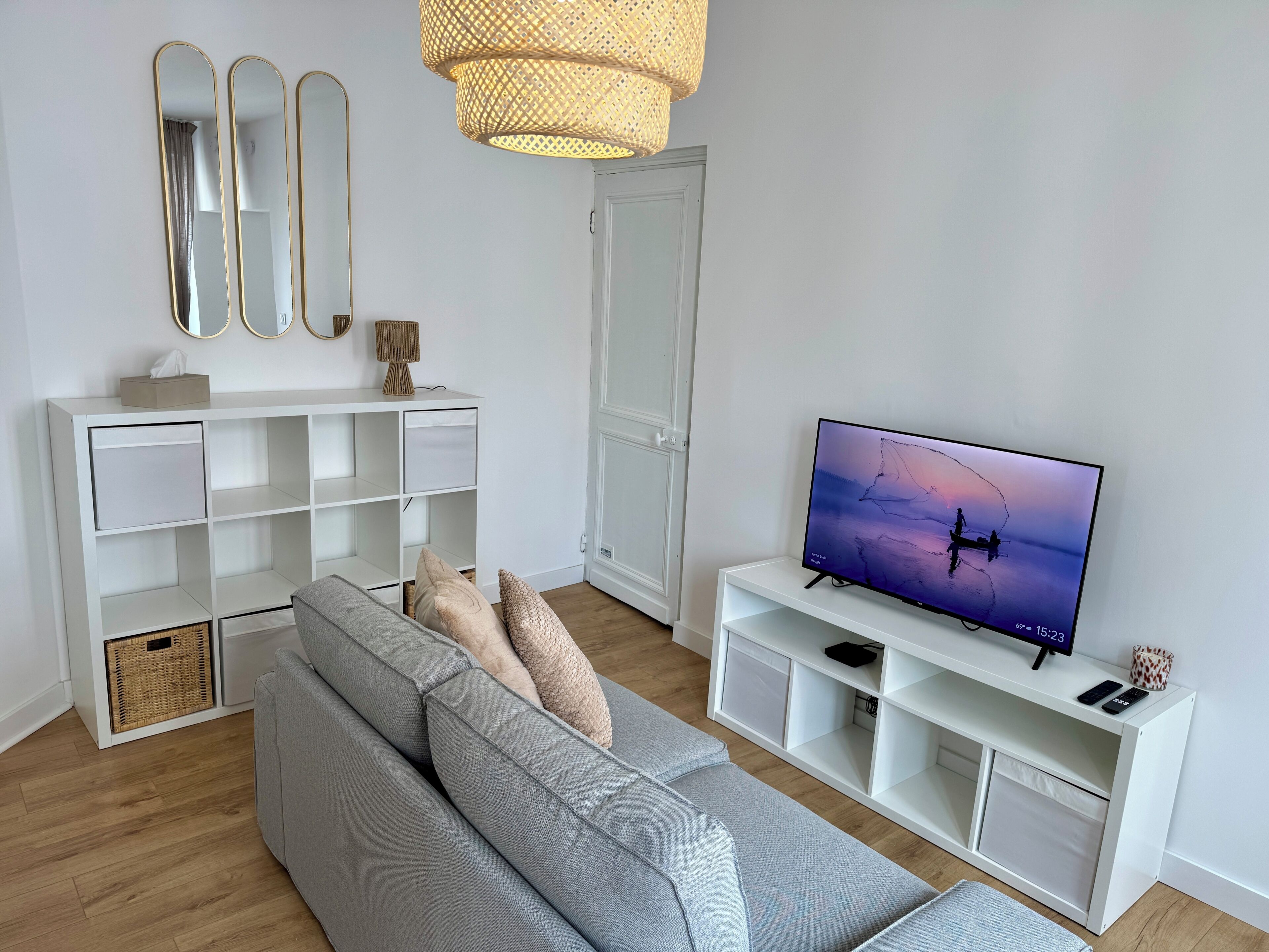 A modern living room with a grey sofa, white shelving, a flat-screen TV, and a woven light fixture.