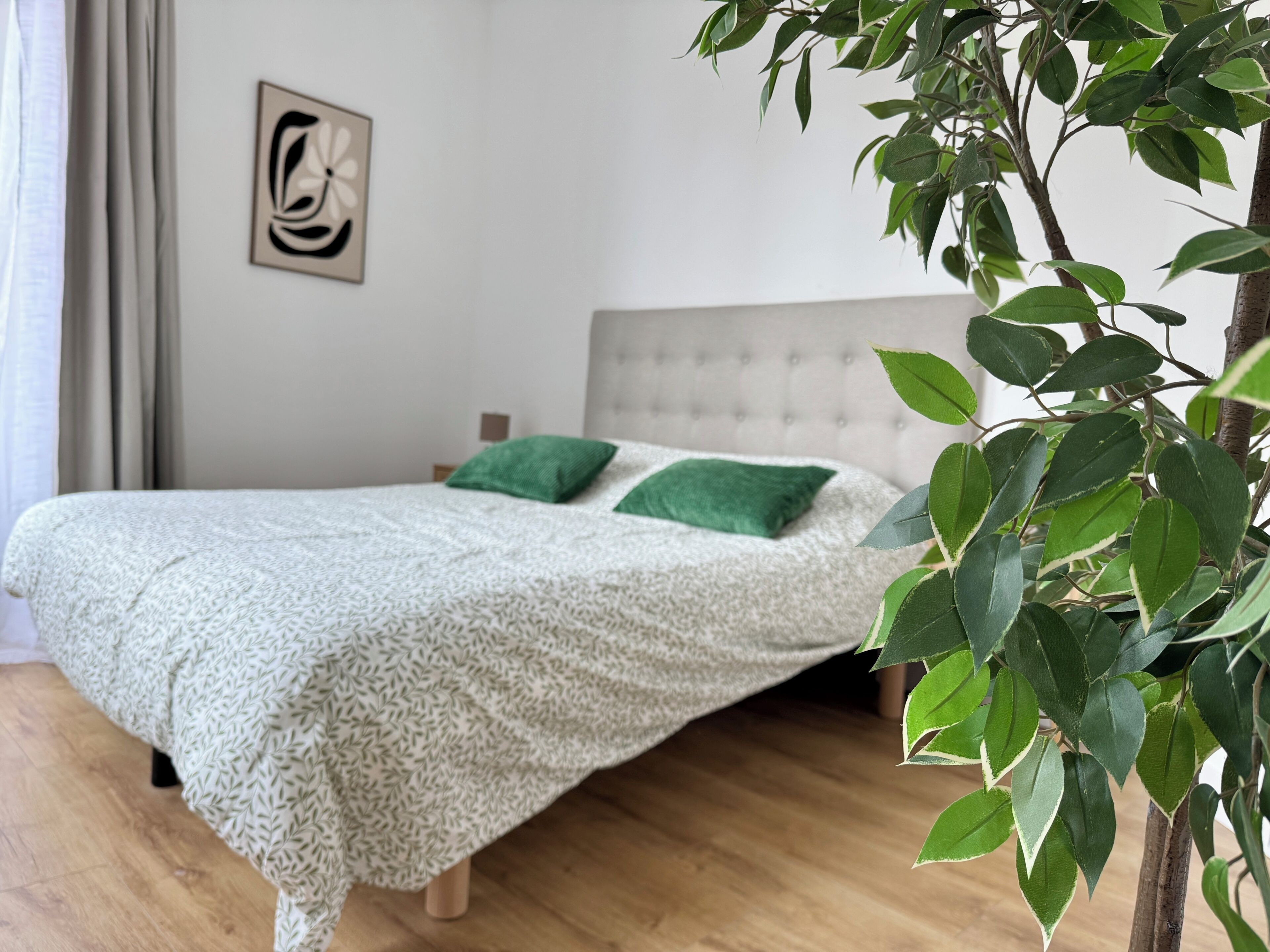 Een slaapkamer met een bed, groene kussens, een gestoffeerd hoofdeinde, een kunstwerk aan de muur en een plant.
