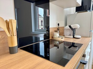A modern kitchen with a black induction cooktop, wooden utensils in a holder, a coffee maker, and a window with a view of a building. - Le Diplomate • T2 rénové au cœur de Douai (Douai)