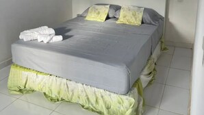Una cama con colchón gris, almohadas blancas y falda de cama verde. Hay una planta en maceta en la pared sobre la cama.