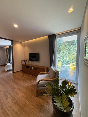 Ein modernes Wohnzimmer mit einem Flachbildfernseher, einer Couch, einem Holztisch und einer großen Glasschiebetür, die auf einen Balkon mit Topfpflanzen führt.