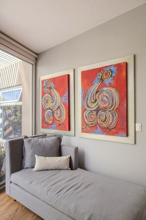 Una habitació amb un sofà gris, dos quadres abstractes a la paret i una finestra amb persianes.