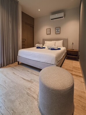A modern bedroom with a bed, a nightstand, a round ottoman, and an air conditioning unit on the wall. - Luxury apartment in Playa del Carmen. (Playa del Carmen)