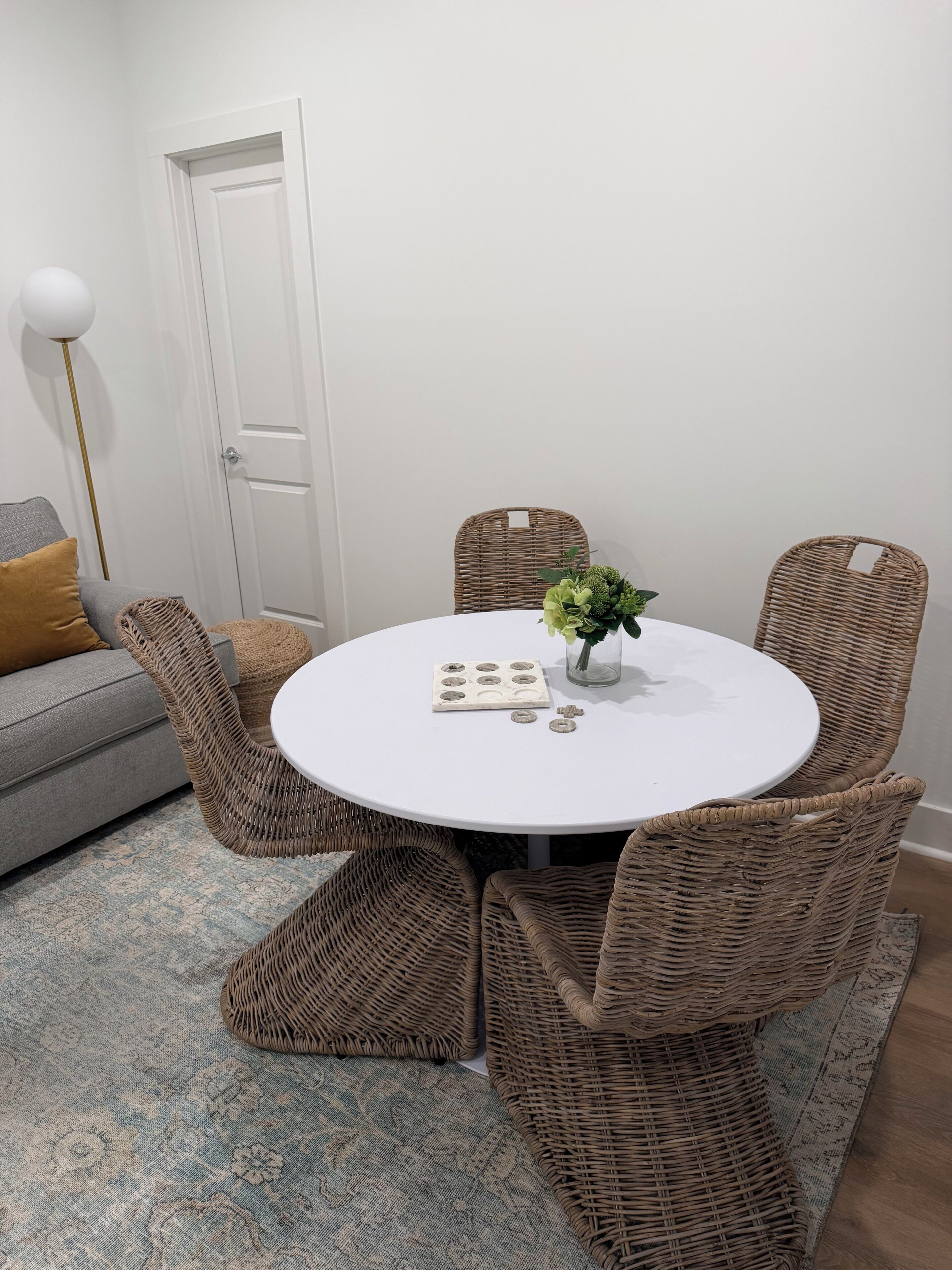 Une table de salle à manger ronde, entourée de chaises en osier, une porte blanche et un canapé gris agrémenté d’un coussin de couleur moutarde.