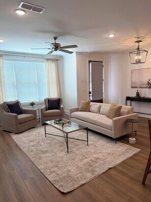 A modern living room with a sofa, two armchairs, a glass coffee table, and a ceiling fan. - Starkville MS Luxury Townhome! Perfect for Gameday! 5 minutes to MSU! (Starkville)
