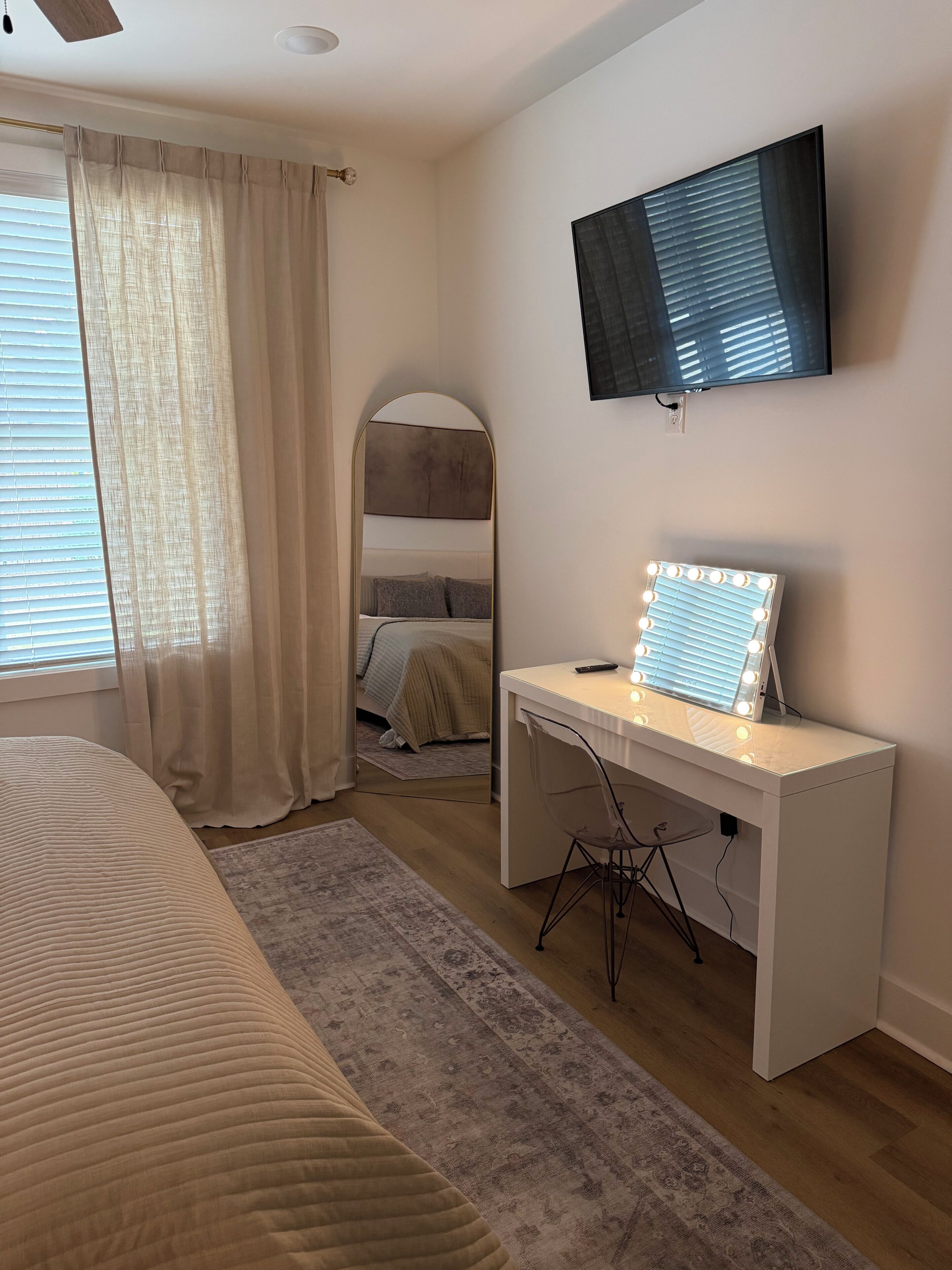 Une chambre à coucher avec un lit, un bureau doté d’un miroir et de lampes, une télévision et une fenêtre avec des rideaux.