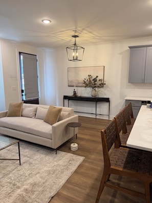 A modern living room with a sofa, a coffee table, a dining table with chairs, a console table with a vase, and a painting on the wall. - Starkville MS Luxury Townhome! Perfect for Gameday! 5 minutes to MSU! (Starkville)