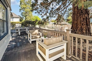 Una terraza de madera con mobiliario exterior, barandilla blanca y un árbol grande.