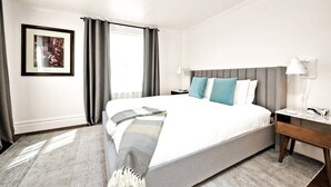 Una camera d'albergo con un letto grande, un comodino con una lampada, un quadro appeso al muro e una finestra con tende.