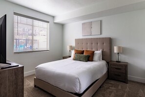 A bedroom with a large bed, two bedside tables, a television on a stand, and a window with blinds. - Landing | Beautiful 1BD, Gym (Draper)