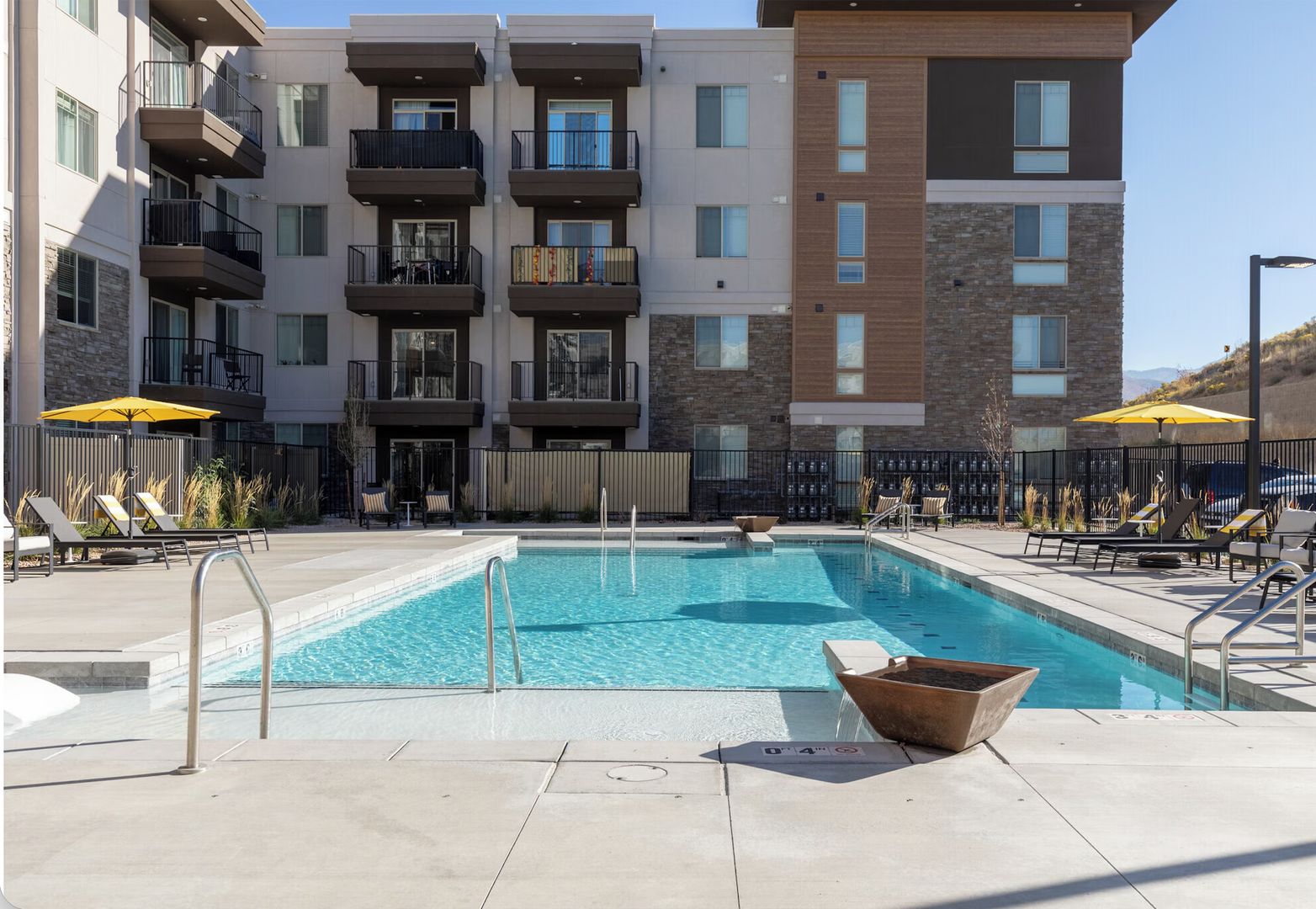 A swimming pool with lounge chairs and umbrellas, surrounded by a multi-story building with balconies.