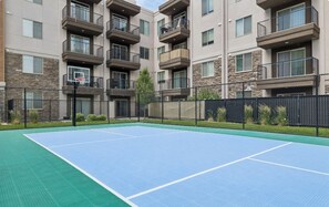 A tennis court with a basketball hoop, surrounded by a fence and residential buildings. - Landing | Incredible 1BD, Gym (Draper)
