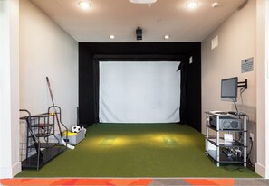 Ein Raum mit grünem Kunstrasenteppich, einer weißen Leinwand an der Wand und einem Computerarbeitsplatz auf der rechten Seite.