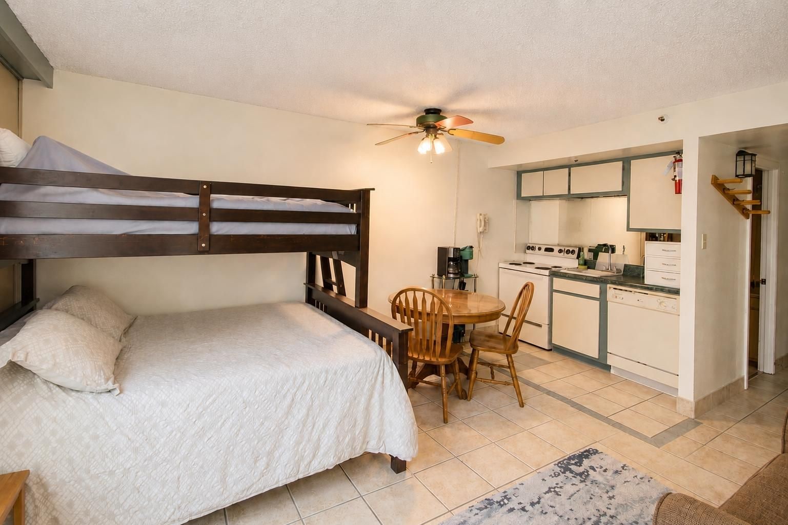 A compact room with a bunk bed, a small dining table with chairs, a kitchenette, and a ceiling fan.