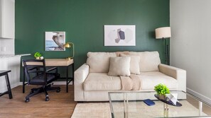 Ein Wohnzimmer mit grüner Wand, einem beigen Sofa, einem Schreibtisch mit Stuhl, einer Lampe und einem Glastisch.