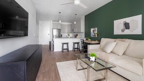 A modern living room with a sofa, a glass coffee table, and a TV. There is a kitchen in the background with white cabinets and a dining area. - Landing | Modern 1BD, Yoga Studio, Gym (Sarasota)