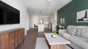 A modern living room with a sofa, coffee table, and TV. There is a kitchen in the background with cabinets and a refrigerator. - Landing | Stunning 1BD, Gym, Yoga Studio (Sarasota)