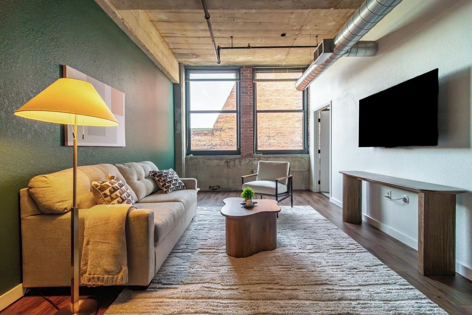A living room with a sofa, armchair, coffee table, and a lamp. There is a TV mounted on the wall and a window with a view of a brick building.