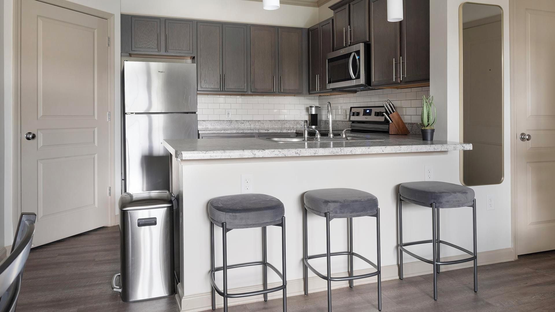 A kitchen with a stainless steel refrigerator, a microwave, a sink, and a bar area with stools.