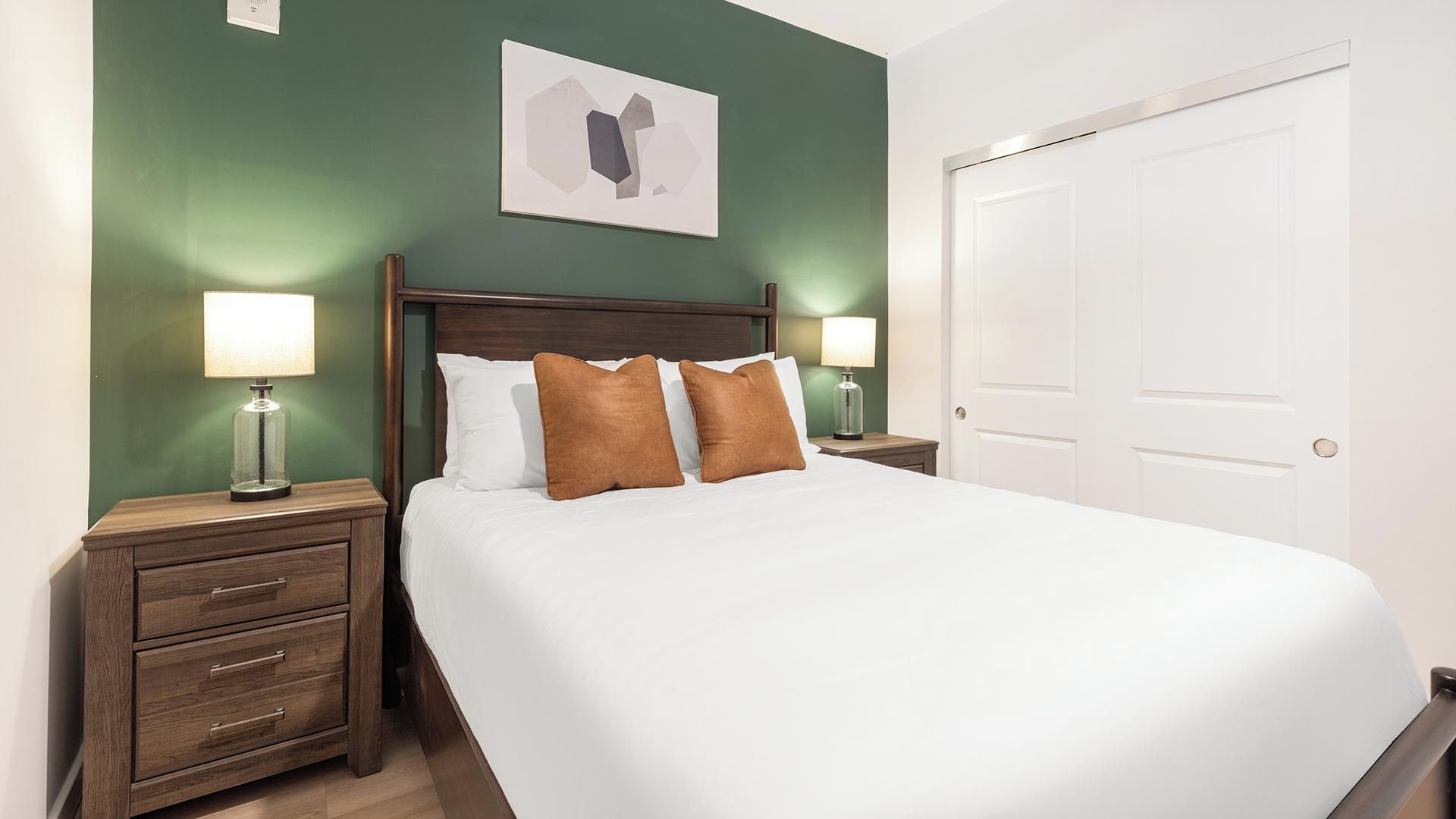 Una cama bien tendida con sábanas blancas y dos almohadas marrones, flanqueada por mesitas de noche de madera con lámparas, en una habitación con un muro verde y una puerta blanca.