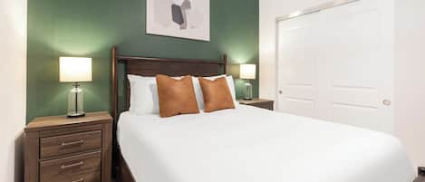 Una cama bien tendida con sábanas blancas y dos almohadas marrones, flanqueada por mesitas de noche de madera con lámparas, en una habitación con un muro verde y una puerta blanca.