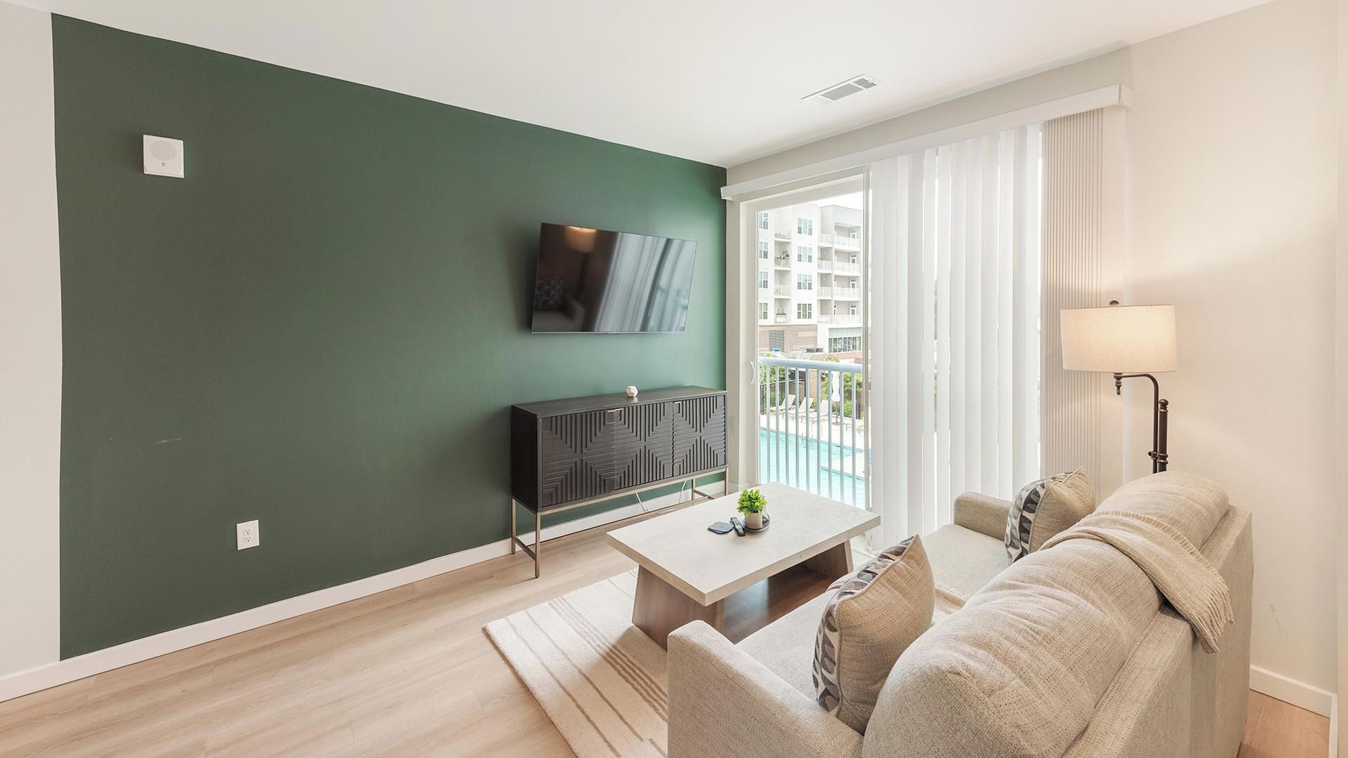 Een moderne woonkamer met een groene accentmuur, een flatscreen televisie, een bank, een salontafel en uitzicht op een balkon met een zwembad.