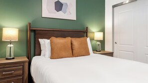 Ein ordentlich gemachtes Bett mit weißen und braunen Kissen, daneben zwei Nachttische mit Nachttischlampen, an einer grünen Wand mit einem gerahmten abstrakten Kunstwerk darüber.